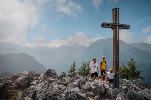 Drei Wanderer stehen an einem Holzkreuz auf einem Berggipfel mit weitem Blick in die Ferne.