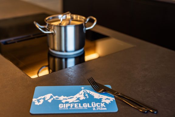 Die Küche bietet ein Induktionskochfeld und ausreichend Töpfe und Pfannen und Geschirr für gesellige Kochabende Silberner Kochtopf auf einem Kochfeld, daneben ein blaues Platzdeckchen und Besteck.