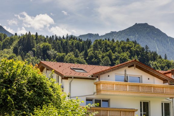Der Neubau aus dem Jahr 2025 liegt erhöht im Berchtesgadener Ortsteil Mitterbach Zweifamilienhaus mit Holzbalustrade, umgeben von grünen Bäumen und Bergen im Hintergrund.