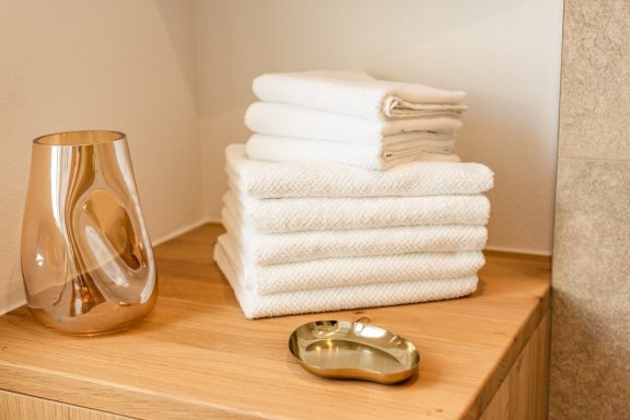 Sie erhalten pro Person einen Satz flauschige Handtücher Gestapelte weiße Handtücher auf einem Holzregal neben einer Vase und einer kleinen Schale.