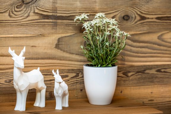 Alpine Details im Gipfelglück Zwei weiße Tierfiguren neben einem Topf mit weißen Blumen auf einer Holzoberfläche.