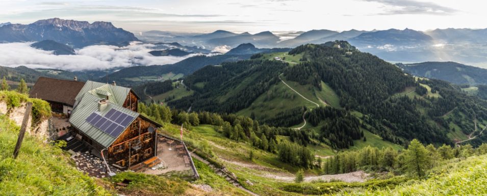 Berglandschaft mit einer Hütte, umgeben von Wiesen und Bergen, im Morgengrauen.