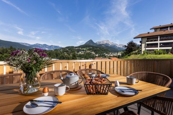Frühstück mit Blick auf die Altstadt von Berchtesgaden vor der Kulisse des Unterbergmassivs Frühstückstisch auf einer Terrasse mit Blumen und Blick auf Berge und blaue Himmel.