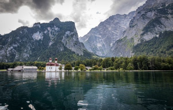 Berglandschaft mit einem See und einer kleinen, historischen Fabrik im Vordergrund.