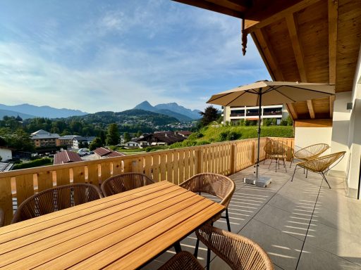 Der Balkon lädt bei Abendsonne zum Entspannen ein Balkon mit Holzterrasse, Blick auf Berge und Häuser unter blauem Himmel.